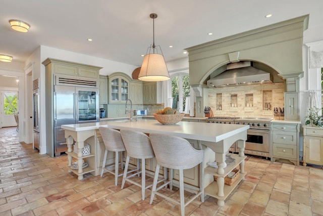 kitchen featuring high quality appliances, a center island with sink, wall chimney exhaust hood, tasteful backsplash, and decorative light fixtures