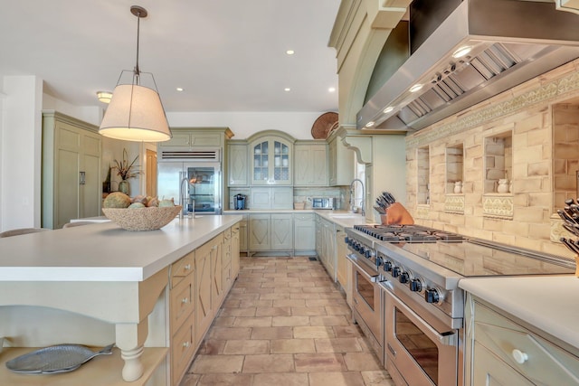 kitchen with pendant lighting, high quality appliances, wall chimney exhaust hood, decorative backsplash, and an island with sink