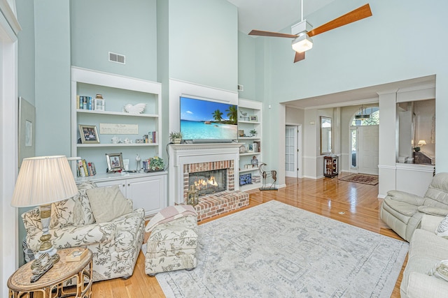 living area with a ceiling fan, visible vents, a brick fireplace, wood finished floors, and a high ceiling