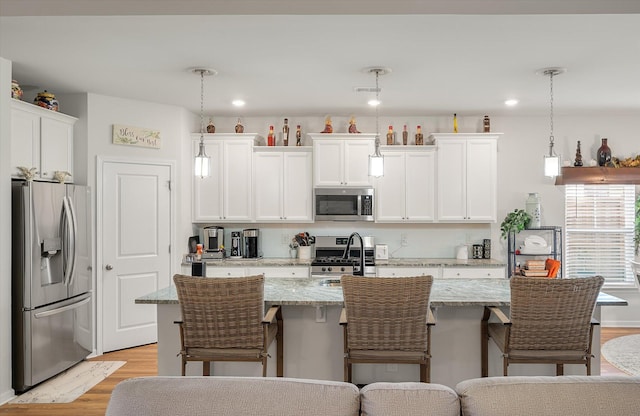 kitchen featuring stainless steel appliances, decorative light fixtures, white cabinetry, an island with sink, and light stone countertops