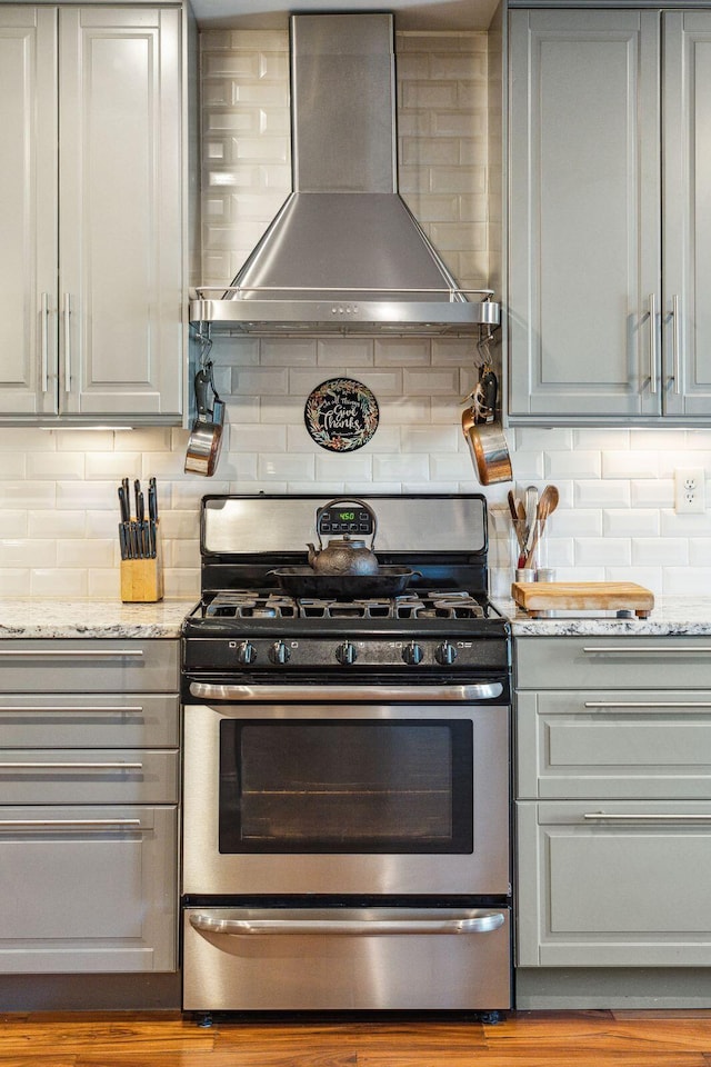 kitchen with light stone counters, tasteful backsplash, gray cabinets, stainless steel gas stove, and wall chimney range hood