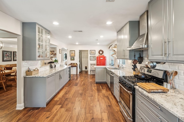 kitchen with gray cabinetry, appliances with stainless steel finishes, wall chimney exhaust hood, dark wood finished floors, and glass insert cabinets