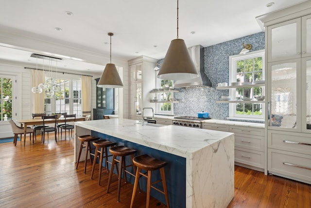 kitchen with wall chimney range hood, sink, light stone counters, an island with sink, and decorative light fixtures