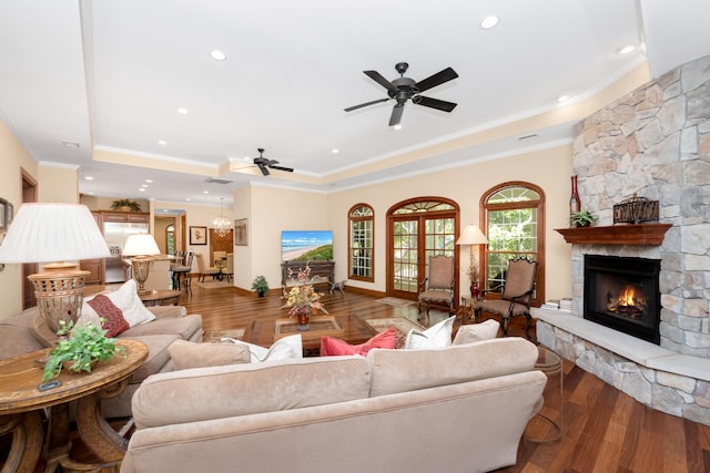 living area with ornamental molding, a stone fireplace, a tray ceiling, and wood finished floors