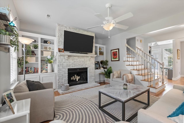 living area with stairs, a brick fireplace, ceiling fan, and wood finished floors