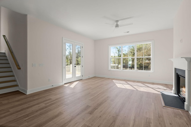 unfurnished living room featuring a glass covered fireplace, ceiling fan, stairway, french doors, and light wood-type flooring