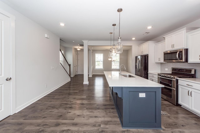 kitchen with an island with sink, a sink, decorative columns, appliances with stainless steel finishes, and white cabinetry