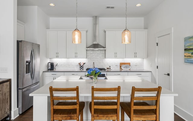 kitchen featuring decorative light fixtures, a center island with sink, stainless steel fridge with ice dispenser, wall chimney range hood, and white cabinets