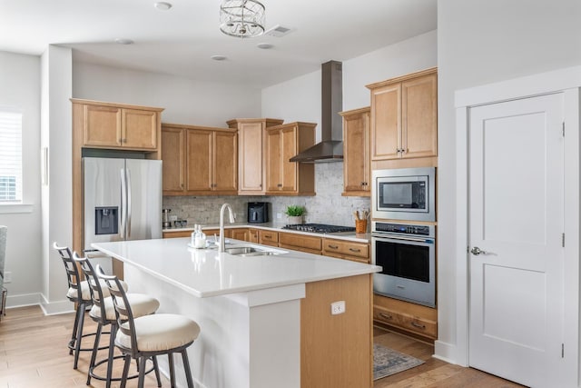 kitchen with a center island with sink, appliances with stainless steel finishes, wall chimney exhaust hood, light hardwood / wood-style flooring, and sink