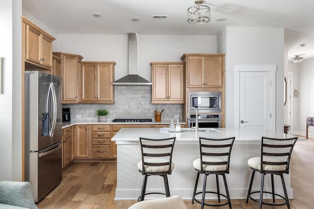 kitchen featuring hardwood / wood-style floors, wall chimney exhaust hood, stainless steel appliances, backsplash, and a center island with sink