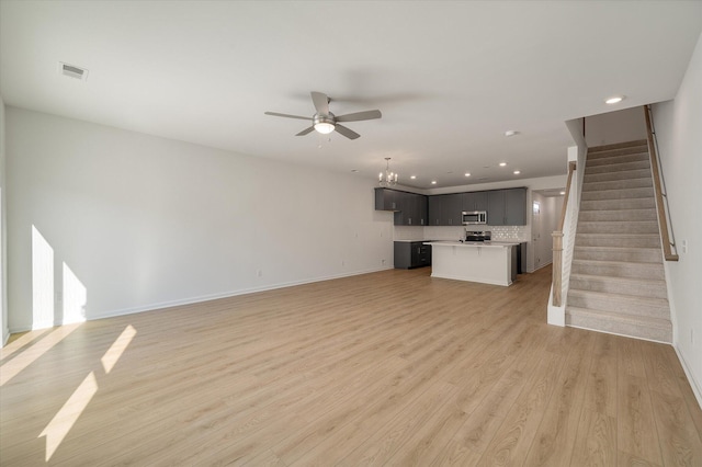 unfurnished living room with ceiling fan with notable chandelier, visible vents, baseboards, stairs, and light wood finished floors