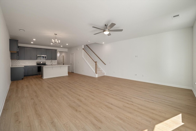 unfurnished living room featuring stairs, light wood finished floors, visible vents, baseboards, and ceiling fan with notable chandelier