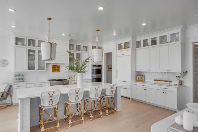 kitchen featuring decorative light fixtures, stainless steel double oven, an island with sink, and white cabinetry