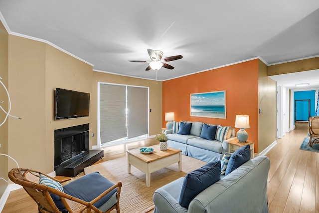 living area featuring a ceiling fan, baseboards, light wood finished floors, a tiled fireplace, and crown molding