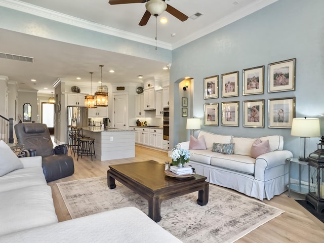 living area featuring light wood finished floors, visible vents, crown molding, ceiling fan with notable chandelier, and arched walkways