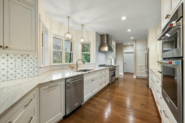 kitchen featuring decorative light fixtures, light stone countertops, stainless steel appliances, wall chimney range hood, and a sink