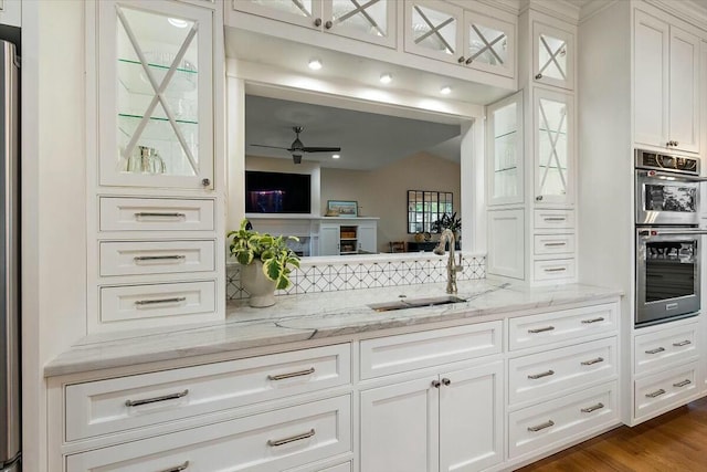 kitchen with stainless steel double oven, a sink, white cabinets, backsplash, and light stone countertops