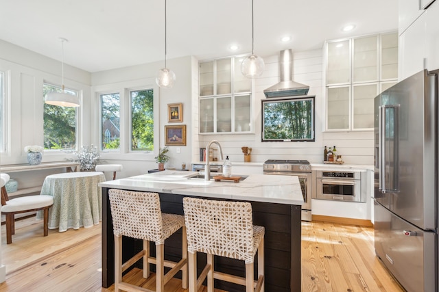 kitchen featuring stainless steel appliances, a center island with sink, wall chimney range hood, and decorative light fixtures