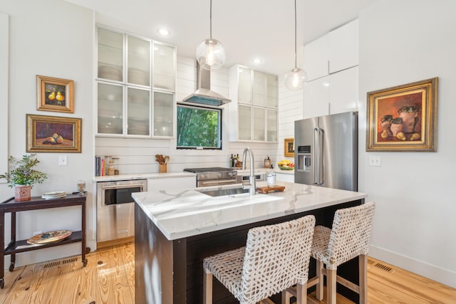 kitchen featuring pendant lighting, appliances with stainless steel finishes, an island with sink, white cabinets, and wall chimney exhaust hood