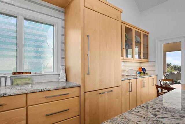 kitchen with tasteful backsplash, glass insert cabinets, light stone counters, and light brown cabinetry