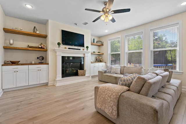 living area featuring light wood-style flooring, a glass covered fireplace, a ceiling fan, and visible vents
