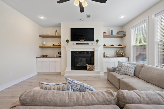 living room with a glass covered fireplace, baseboards, light wood-type flooring, a ceiling fan, and visible vents