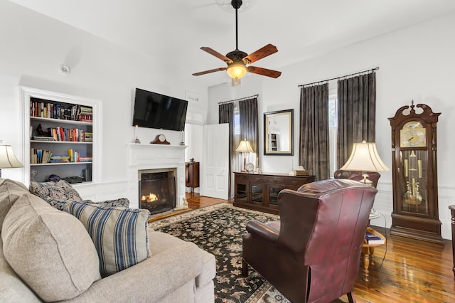 living area with built in features, wood-type flooring, a warm lit fireplace, and a ceiling fan