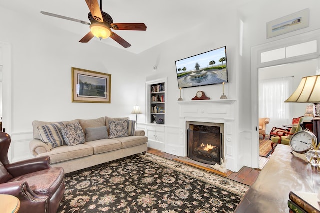 living area featuring built in features, a warm lit fireplace, a ceiling fan, and wood finished floors