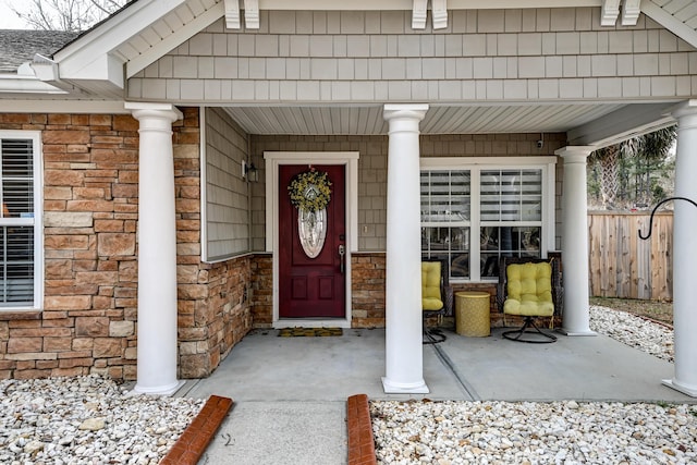 doorway to property with stone siding, a porch, a shingled roof, and fence