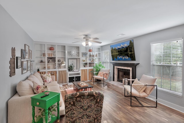 living area featuring built in features, a brick fireplace, ceiling fan, wood finished floors, and baseboards