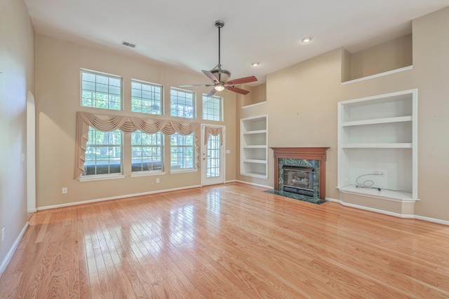 unfurnished living room featuring a high end fireplace, a towering ceiling, built in shelves, ceiling fan, and light hardwood / wood-style flooring