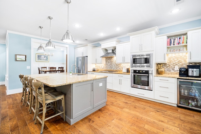 kitchen with beverage cooler, wall chimney exhaust hood, appliances with stainless steel finishes, a kitchen island with sink, and white cabinetry