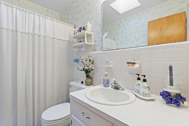 full bath with wallpapered walls, a skylight, toilet, a wainscoted wall, and vanity