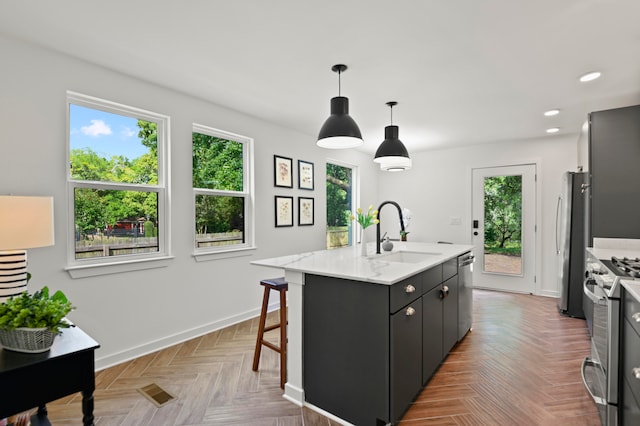 kitchen with sink, stainless steel appliances, light parquet floors, decorative light fixtures, and a center island with sink