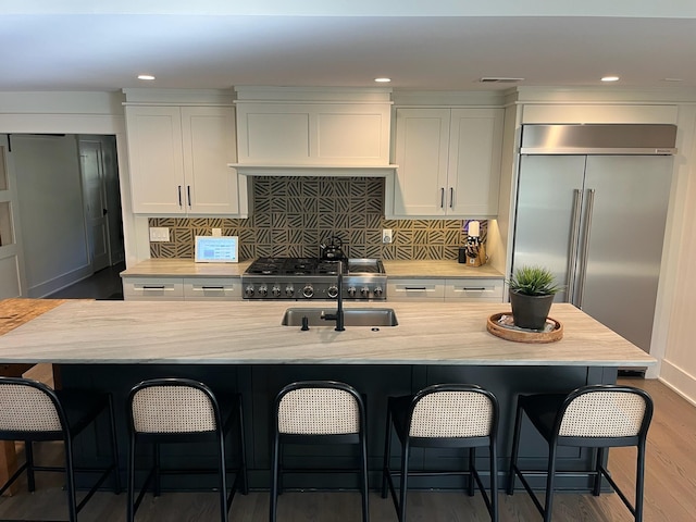 kitchen with built in refrigerator, an island with sink, wood finished floors, white cabinets, and decorative backsplash