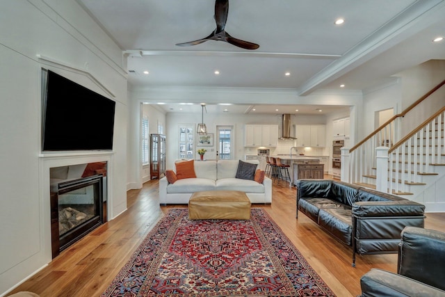 living room featuring a glass covered fireplace, stairs, light wood-type flooring, and ceiling fan