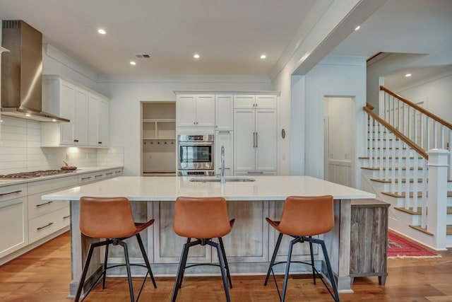 kitchen with a sink, light wood finished floors, appliances with stainless steel finishes, and wall chimney range hood