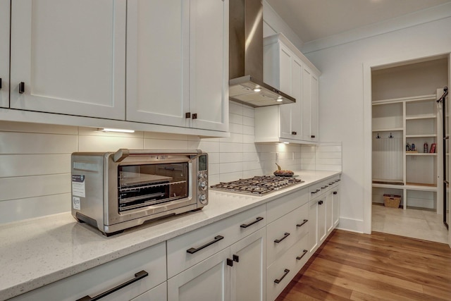 kitchen featuring light wood-type flooring, white cabinets, stainless steel gas stovetop, wall chimney range hood, and tasteful backsplash