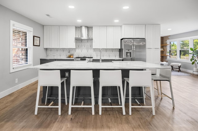 kitchen with stainless steel refrigerator with ice dispenser, wall chimney exhaust hood, baseboards, backsplash, and a kitchen bar