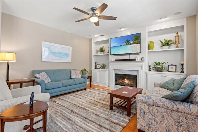 living room featuring built in features, wood finished floors, a fireplace with flush hearth, and ceiling fan