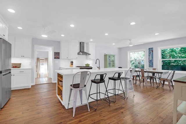 kitchen with wall chimney range hood, ceiling fan, light wood-style floors, freestanding refrigerator, and a center island with sink
