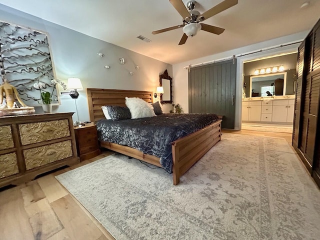 bedroom featuring light hardwood / wood-style floors, ensuite bathroom, a barn door, and ceiling fan