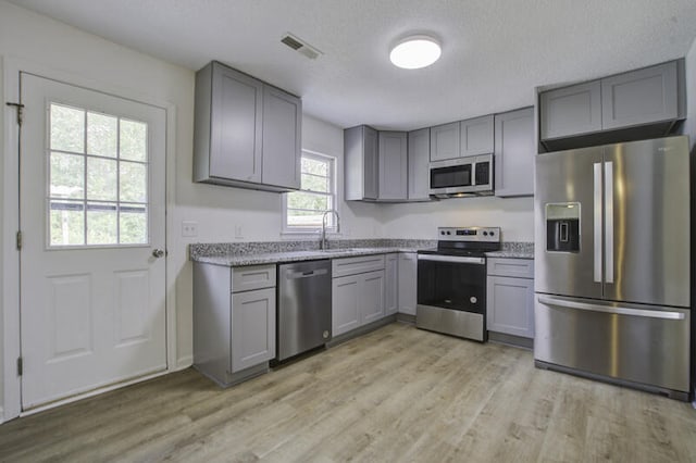 kitchen with light wood-type flooring, gray cabinetry, a textured ceiling, stainless steel appliances, and light stone countertops