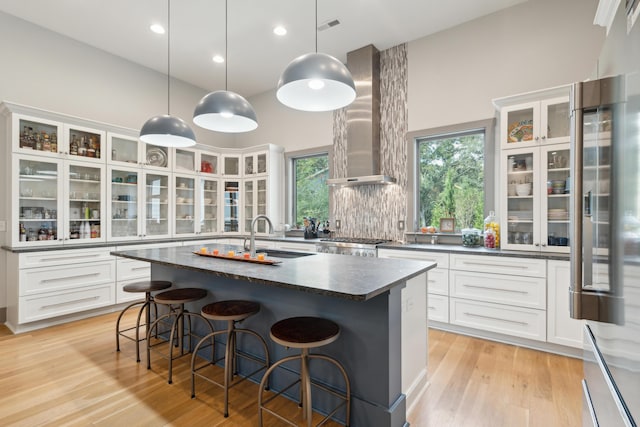 kitchen with white cabinetry, an island with sink, hanging light fixtures, and wall chimney range hood