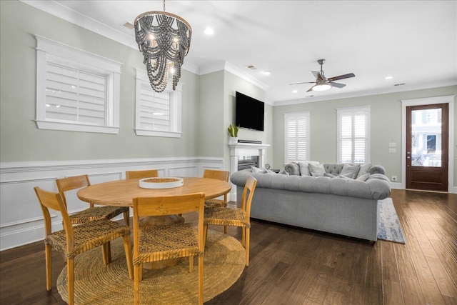 dining area with a wainscoted wall, ceiling fan with notable chandelier, crown molding, and dark wood-style flooring