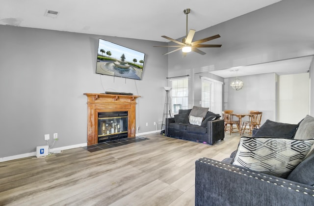 living room featuring wood finished floors, visible vents, baseboards, a fireplace with flush hearth, and ceiling fan with notable chandelier