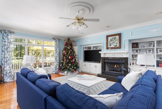 living room with built in features, a ceiling fan, light wood-style flooring, a lit fireplace, and crown molding