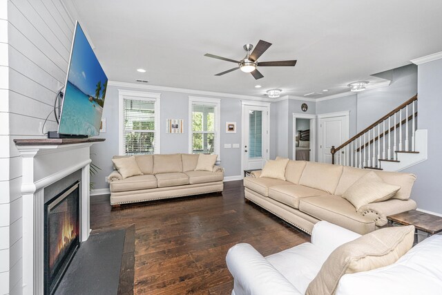 living room featuring wood-type flooring, a glass covered fireplace, ornamental molding, a ceiling fan, and stairs