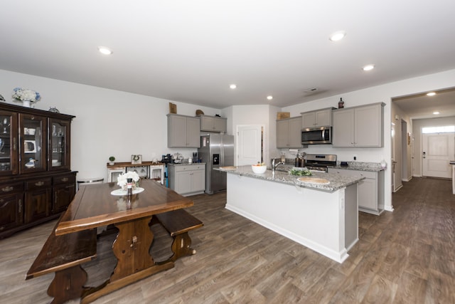 kitchen with light stone countertops, stainless steel appliances, a center island with sink, dark wood-type flooring, and gray cabinetry