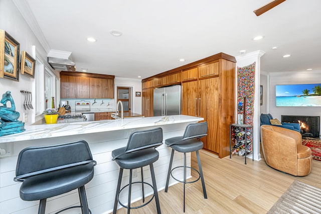 kitchen featuring high end refrigerator, a breakfast bar area, light stone countertops, a lit fireplace, and crown molding